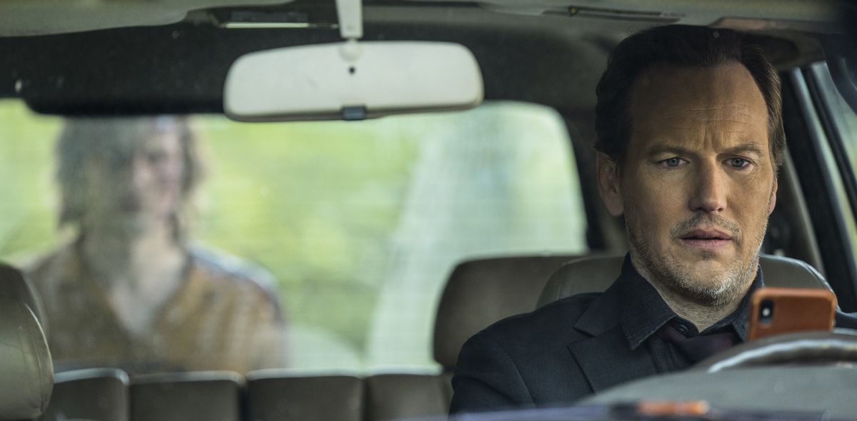 Josh (Patrick Wilson), homme d'âge moyen à l'air perturbé, est assis sur le siège du conducteur d'une voiture et regarde son téléphone portable tandis qu'une silhouette humanoïde floue à la peau grise et au visage apparemment ensanglanté se profile devant le pare-brise arrière dans Insidious : La Porte Rouge