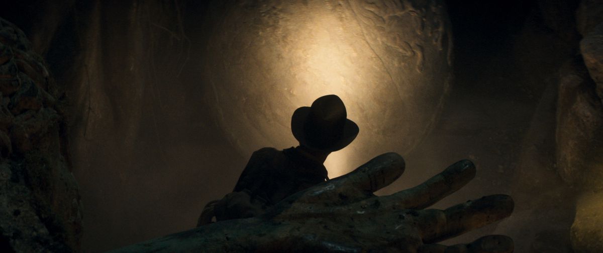 Silhouette d'Indiana Jones explorant une tombe, la main d'un cadavre est au premier plan, également ombragée.