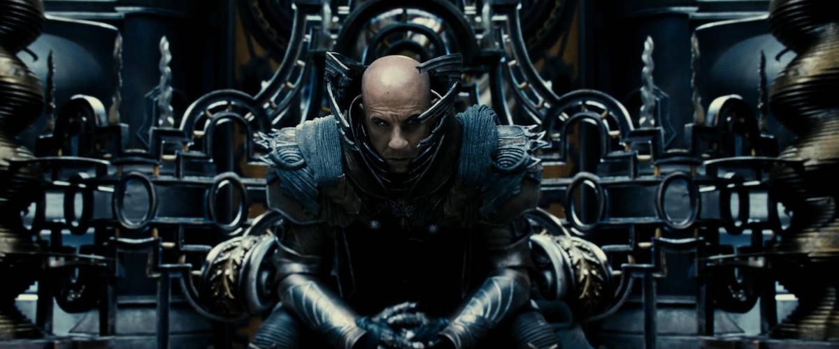Un homme chauve (Vin Diesel) est assis sur un trône, vêtu d'une armure métallique élaborée et entouré d'une technologie élaborée sans objectif clair dans Les Chroniques de Riddick.