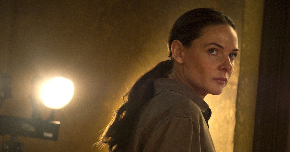 Ilsa Faust (Rebecca Ferguson), les cheveux tirés en queue de cheval, se tient dans une pièce jaune-brun quelconque avec une lumière vive derrière elle dans Mission : Impossible - Dead Reckoning Part One
