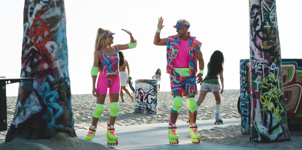 Barbie (Margot Robbie) et Ken (Ryan Gosling), tous deux vêtus de tenues de patinage au néon à motifs criards et de genouillères et rollers jaune fluo incroyablement brillants, se tiennent devant une plage entre deux arbres couverts de graffitis et entrent pour un high five in le film d'action en direct Barbie de 2023