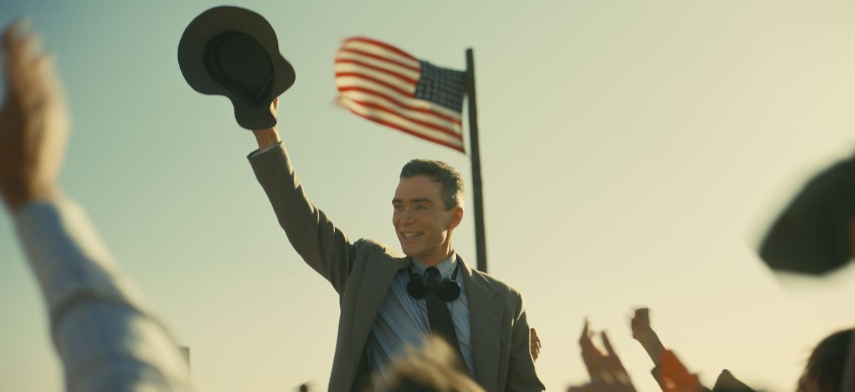 J. Robert Oppenheimer (Cillian Murphy) sourit et agite son chapeau devant une foule en plein air en liesse alors qu'il se tient sous un drapeau américain dans Oppenheimer de Christopher Nolan