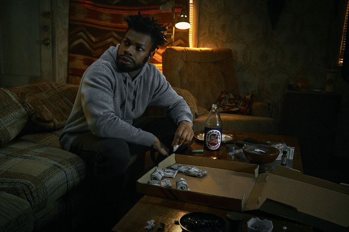 John Boyega dans le rôle de Fontaine assis sur un canapé devant une boîte à pizza remplie de rouleaux de billets d'un dollar dans They Cloned Tyrone.