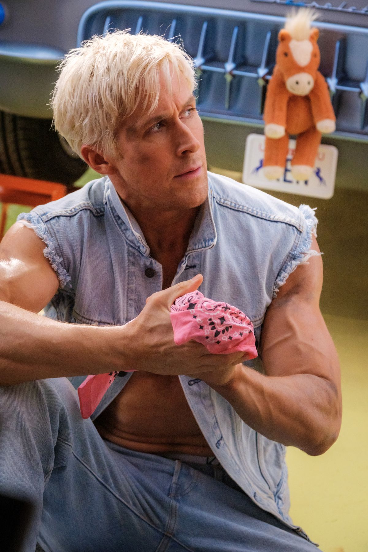 Ryan Gosling dans le rôle de Ken dans un gilet en jean et un jean, un petit bandana rose dans les mains.  Il est assis par terre.  Derrière lui, il y a un camion, avec un cheval en peluche attaché à l'avant. 