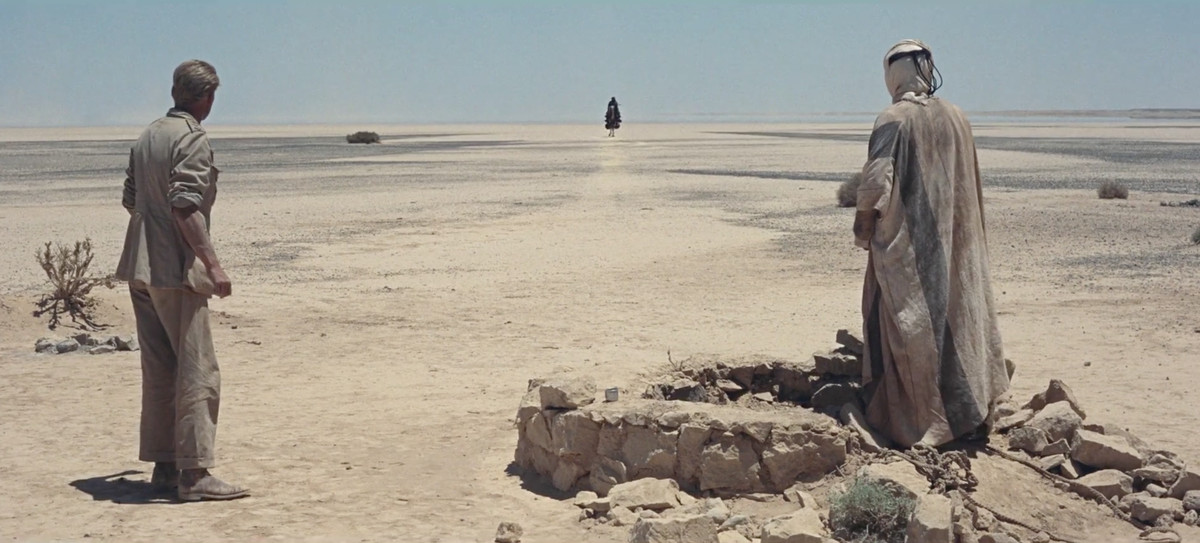 Deux hommes vêtus de vêtements ternes et sales se tiennent dos à la caméra dans un désert aride de la même couleur que leurs vêtements, alors qu'une silhouette solitaire s'approche au loin.