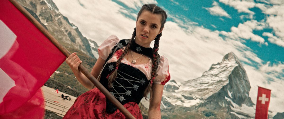 Alice Lucy dans le rôle de Heidi, vêtue d'une robe éclaboussée de sang, tenant un drapeau rouge avec les Alpes suisses en arrière-plan dans Mad Heidi.