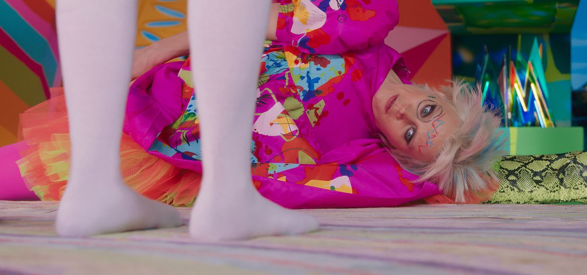 Weird Barbie (Kate McKinnon), une Barbie dans une robe informe, ample et multicolore, avec ses cheveux coupés à différentes longueurs courtes teints en rose pastel et bleu, et avec des gribouillis sur son visage, est allongée sur le sol en regardant les pieds chaussés et sans chaussures de Barbie (Margot Robbie) dans le film d'action en direct Barbie de 2023.