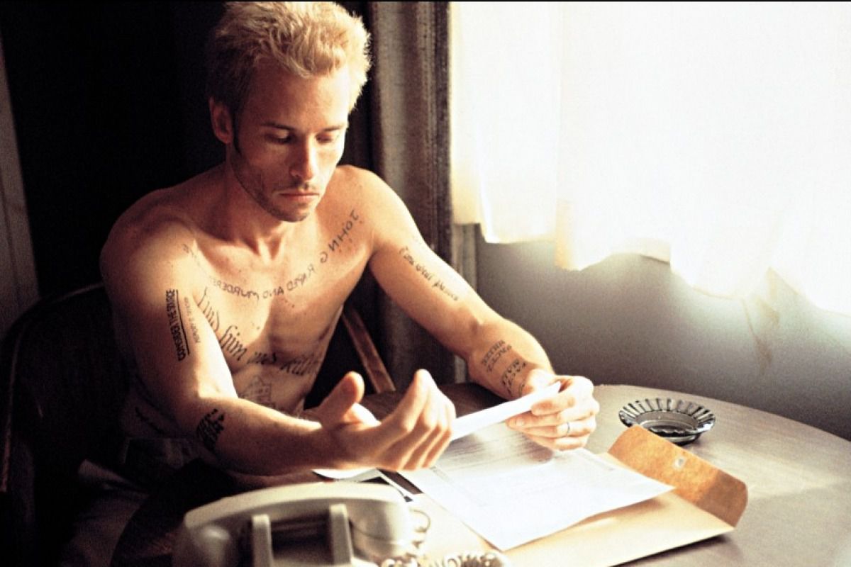 Leonard Shelby (Guy Pearce) est assis torse nu devant une fenêtre à rideaux dans une chambre d'hôtel, couvert de tatouages ​​de rappel et avec quelques papiers devant lui dans Christopher Nolan's Memento.