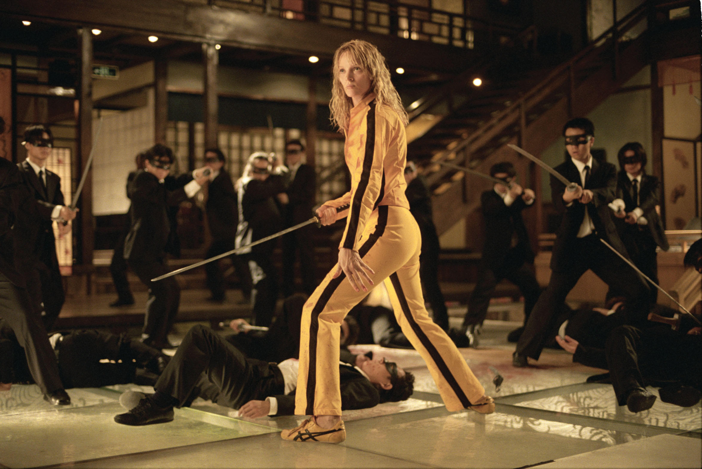 Uma Thurman dans le rôle de The Bride, vêtue d'une combinaison jaune et tenant une épée, est entourée des Crazy 88 Yakuzas dans Kill Bill Vol.  1