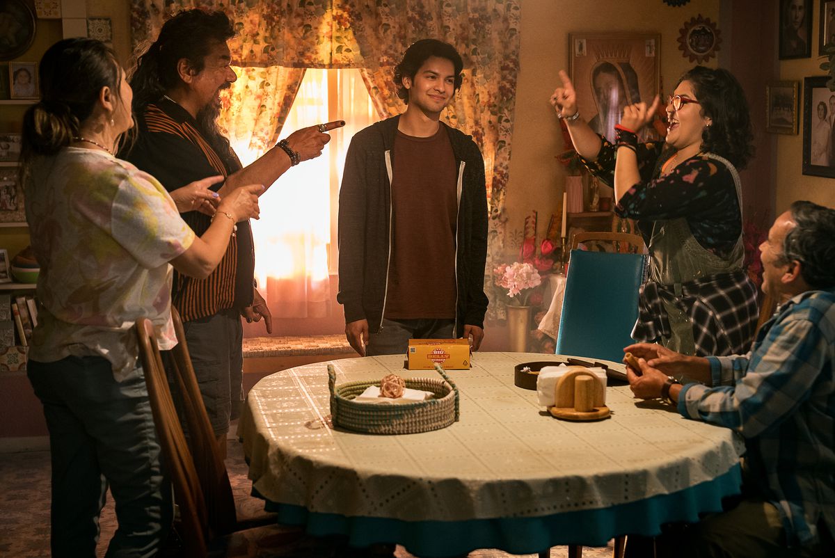 Jaime Reyes se tient penaud derrière la table du dîner familial tandis que toute sa famille le pousse de manière ludique dans le film Blue Beetle