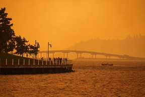 Une personne voyage dans un bateau devant des personnes marchant sur la promenade alors que la fumée du feu de forêt de McDougall Creek recouvre la zone du lac Okanagan, à Kelowna, en Colombie-Britannique, le vendredi 18 août 2023.