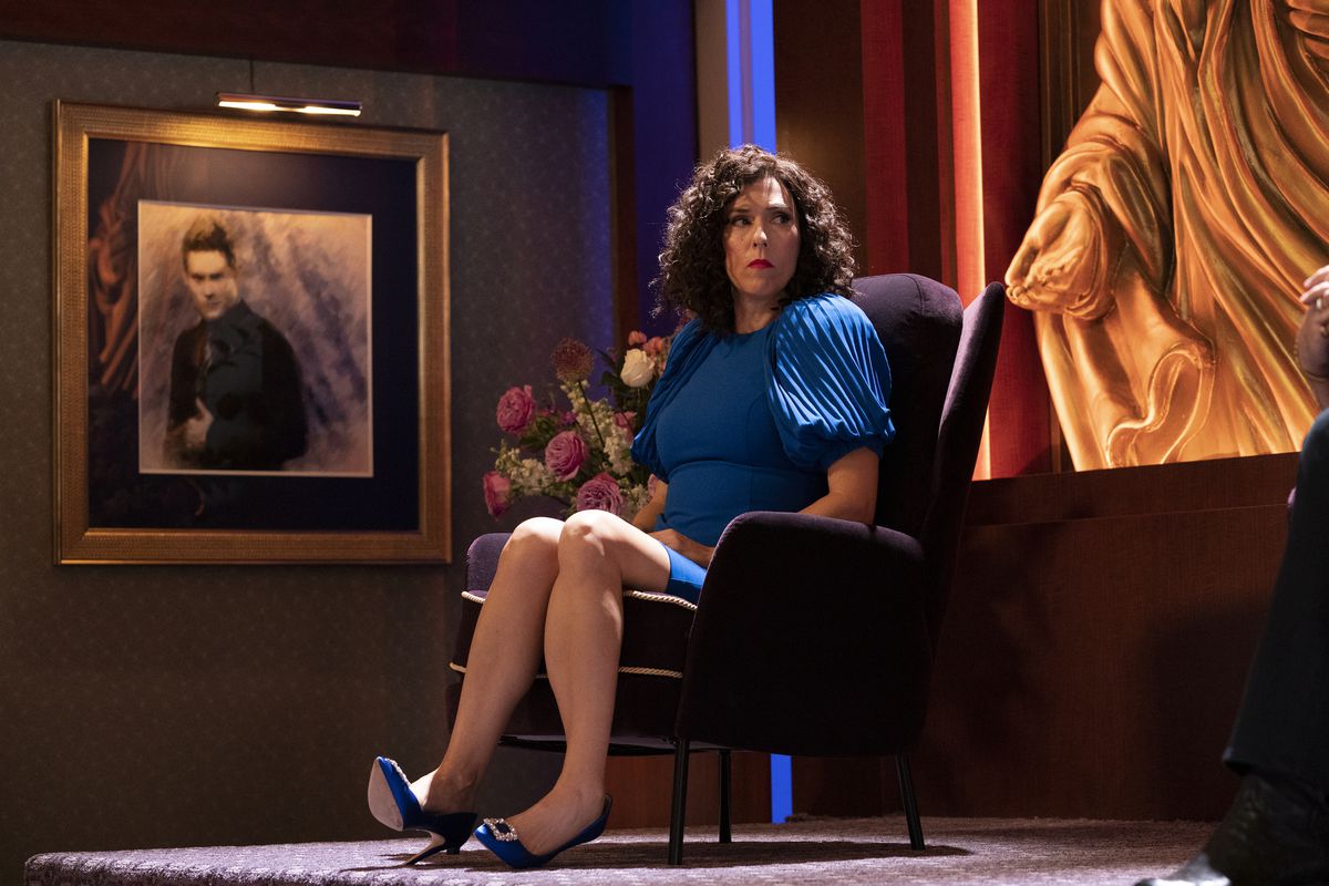 Judi Patterson, vêtue d'une robe bleue avec des talons bleus, est assise sur une chaise violette sur une plate-forme surélevée dans The Righteous Gemstones.  Derrière elle se trouve une statue géante d'elle-même, tandis qu'à droite se trouve un dessin encadré de Kelvin Gemstone (Adam Devine).