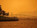 Une personne voyage dans un bateau devant des gens marchant sur la promenade alors que la fumée de l'incendie de forêt de McDougall Creek recouvre la zone du lac Okanagan, à Kelowna, en Colombie-Britannique, le vendredi 18 août 2023.