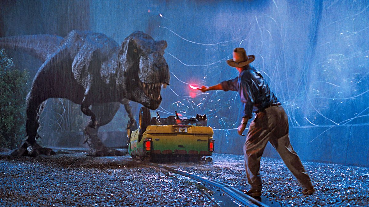 Un homme portant un chapeau marron et une chemise bleue (Sam Neill) agite une fusée éclairante d'urgence allumée pour distraire un Tyrannosaurus Rex debout à côté d'un camion vert et jaune renversé à côté d'une porte métallique cassée.
