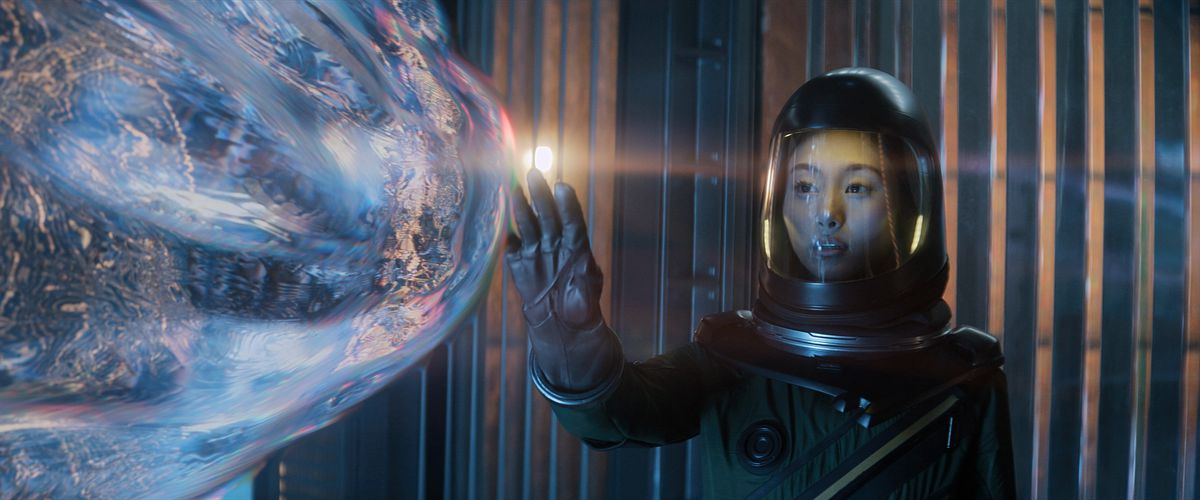 Une femme en combinaison spatiale tend la main pour toucher une goutte réfractant la lumière et venant apparemment vers elle comme si elle était vivante dans la saison 2 d'Invasion d'Apple.