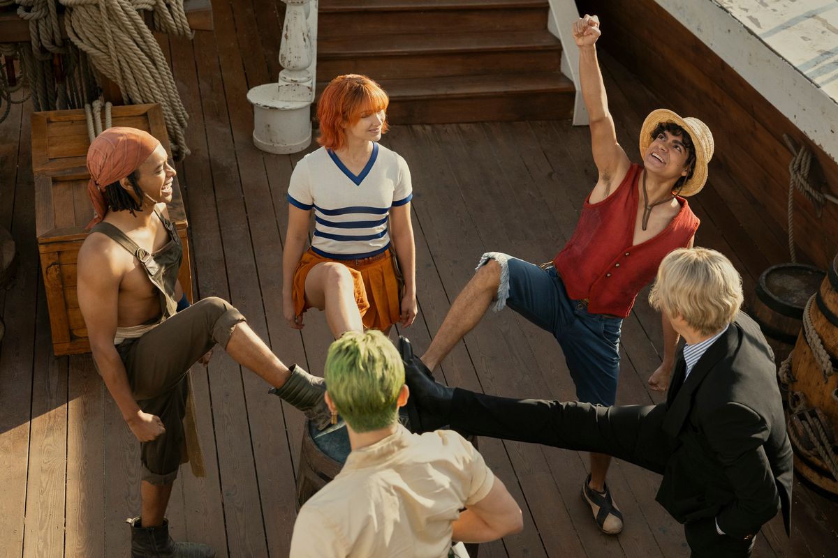 Usopp, Zoro, Nami, Luffy et Sanji se tiennent les jambes levées au centre d'un bateau pirate tandis qu'une équipe applaudit dans One Piece.