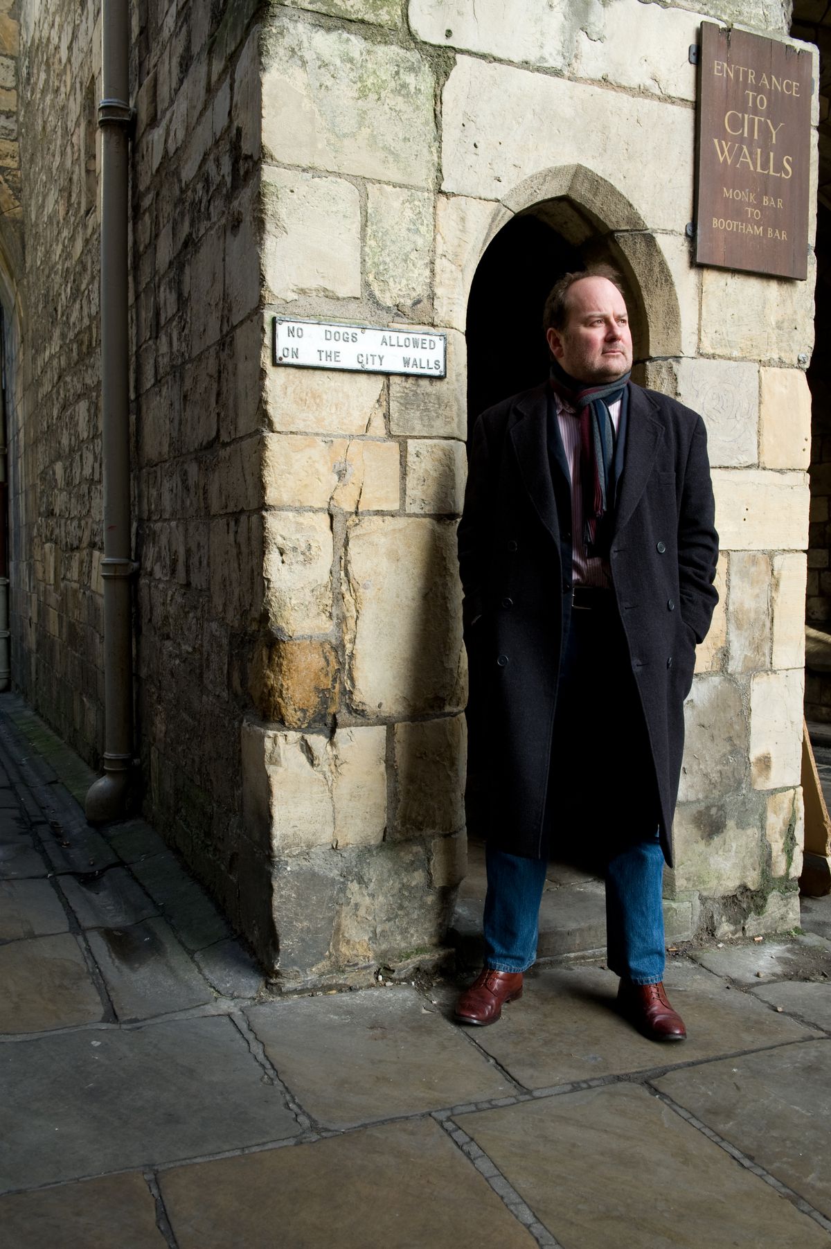 Charles Cecil, en manteau d'hiver et écharpe, se tient devant les anciens murs de la ville de York