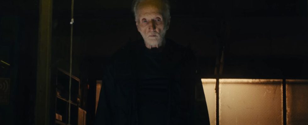 Tobin Bell stands ominously in a warehouse, dressed in black, in Saw X.