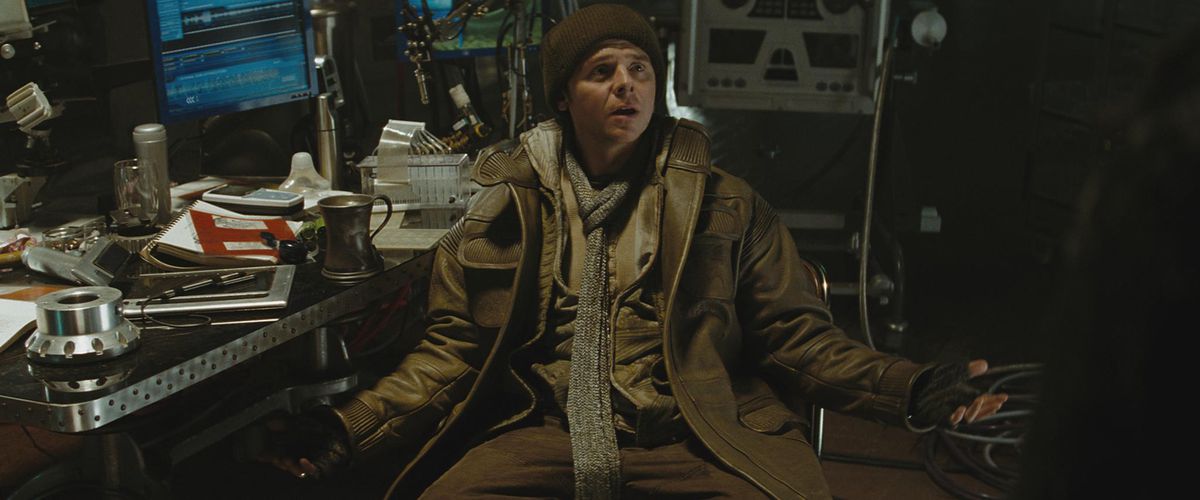 Simon Pegg dans le rôle de Scotty dans Star Trek (2009) écarte les bras dans un geste interrogateur, assis dans des vêtements chauds à un bureau encombré. 