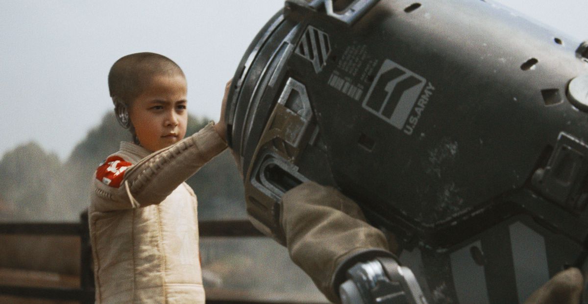 Un jeune enfant androïde au crâne rasé et au crâne partiellement manquant pose sa main sur une machine dans The Creator