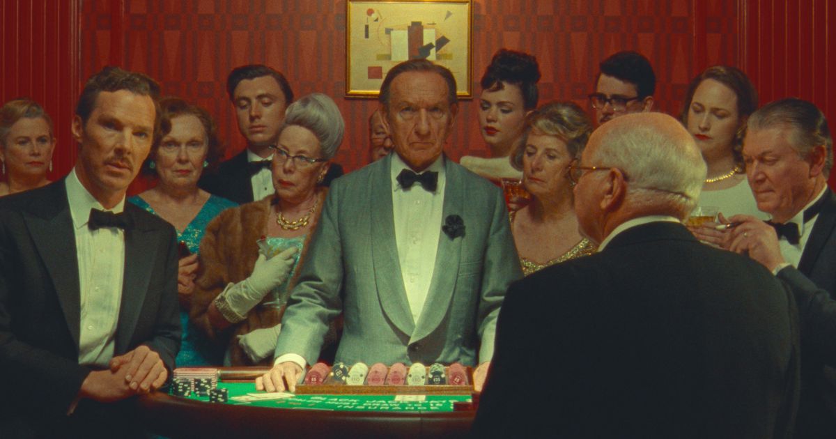 Benedict Cumberbatch (en smoking dans le rôle d'Henry Sugar) et Sir Ben Kingsley (en croupier) regardent la caméra alors qu'ils se tiennent à une table dans un casino, entourés d'une foule curieuse de gens bien habillés, dans le film Netflix de Wes Anderson The Merveilleuse histoire d’Henry Sugar