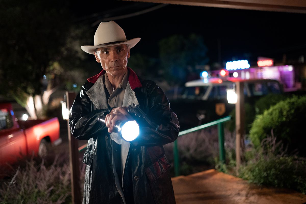 Zahn McClarnon dans le rôle de l'officier Joe Leaphorn lève une lampe de poche et une arme à feu lors d'une nuit de désert avec des néons derrière lui dans Dark Winds d'AMC.
