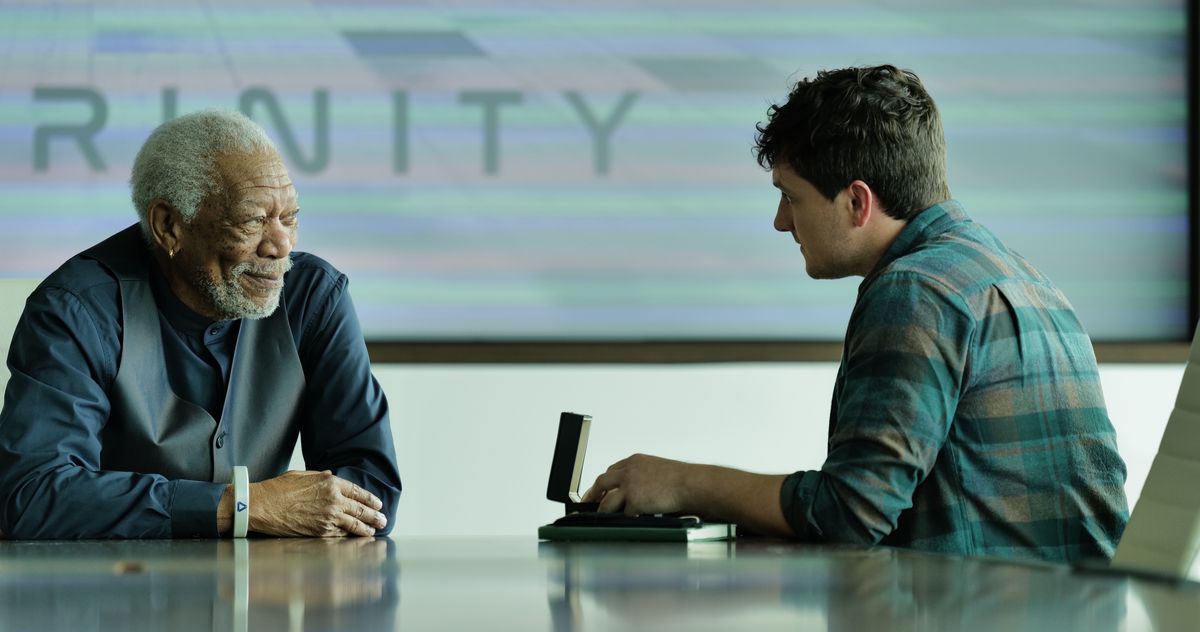 Morgan Freeman, vêtu d'un élégant costume bleu-gris et sans veste, est assis, souriant, devant une table de réunion brillante du journaliste Josh Hutcherson, devant un mur sur lequel est écrit 