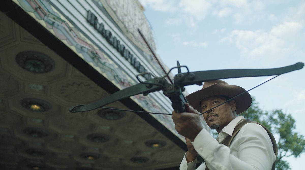 Terrence Howard tient une arbalète devant un chapiteau de cinéma tout en portant un chapeau de cowboy dans Showdown at the Grand.