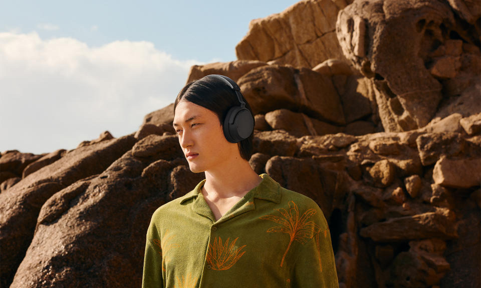 Photo marketing lifestyle pour le casque Sennheiser Accentum Wireless.  Un jeune homme se tient debout sur un terrain rocheux : des rochers saillants derrière lui.  Il porte une chemise boutonnée verte à motifs floraux or/orange.