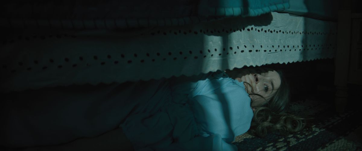 Brynn Adams (Kaitlyn Dever), une jeune femme aux cheveux longs vêtue d'une chemise de nuit bleue, se cache sous son lit dans le noir avec sa main posée sur sa bouche dans le film d'horreur No One Will Save You