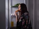 Femme mangeant un éclair devant la porte ouverte du réfrigérateur.