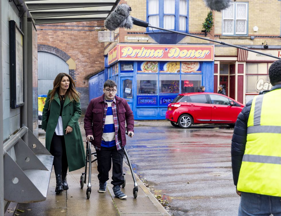 Bobby et Carla dans Coronation Street, dans les coulisses du tournage d'une femme et d'un jeune homme avec une aide à la marche se promenant dans la rue
