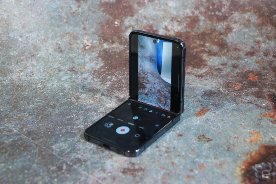 Photo du Samsung Z Flip 5 posé sur une surface qui ressemble à du métal brut avec des taches rouillées.  Le téléphone est partiellement ouvert et la vue de la caméra est visible sur la moitié supérieure de l'écran.