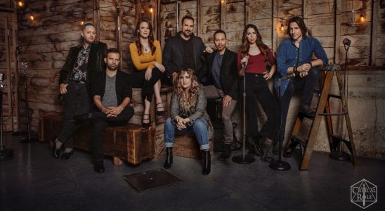 The cast of Critical Role in a rustic wooden building, standing around or sitting on crates with microphones surrounding them