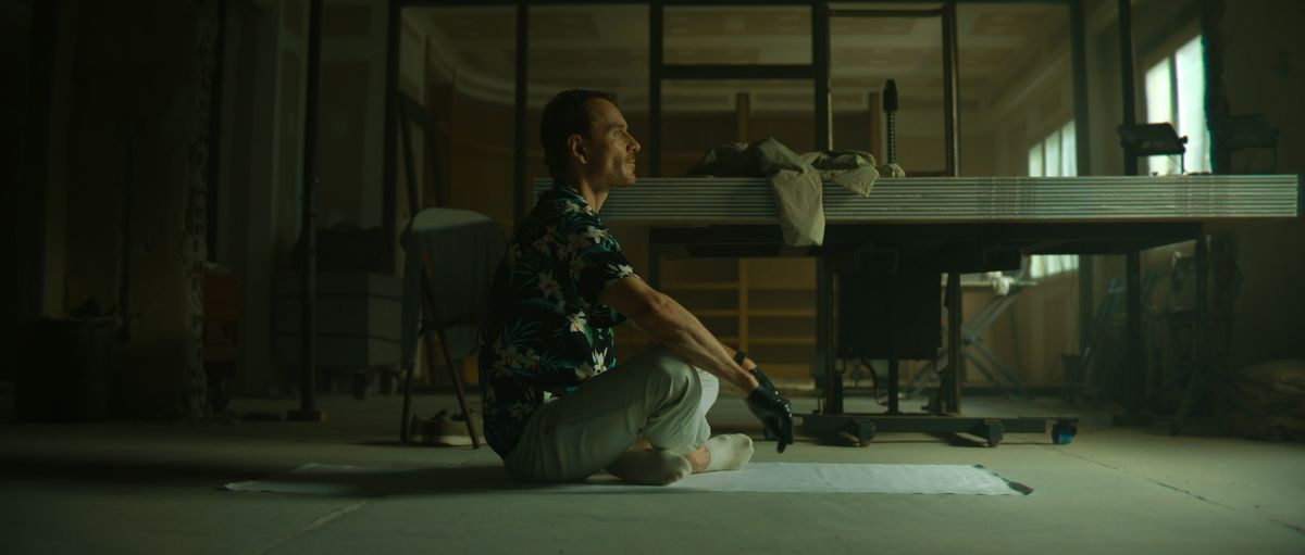 Michael Fassbender dans le rôle de The Killer est assis les jambes croisées sur le sol sur une feuille de plastique
