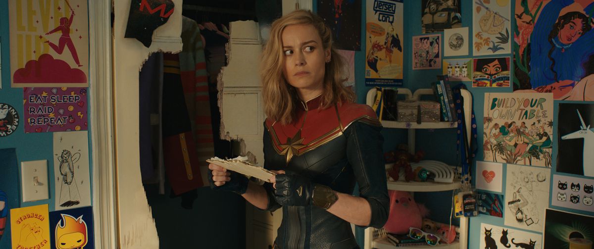 Le capitaine Marvel (Brie Larson) se tient dans la chambre de Kamala Khan à côté d'une porte de placard brisée, regardant autour de lui avec inquiétude toutes les images de dessins animés d'elle-même, dans le film Marvel Cinematic Universe The Marvels.