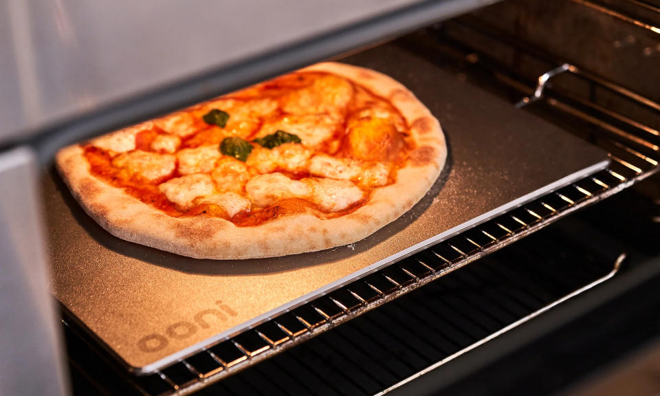 Photo marketing lifestyle de l'Ooni Pizza Steel 13, une plaque en acier inoxydable pour pizza cuite au four.  Une porte de four est ouverte alors que nous voyons une pizza prête à manger sur la surface en acier.