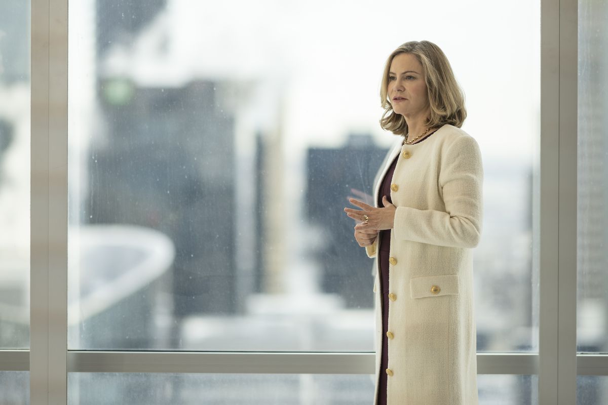 Jennifer Jason Leigh dans le rôle de Lorraine Lyon se tient dans une blouse blanche coûteuse devant la fenêtre d'un bureau dans la saison 5 de Fargo de FX.