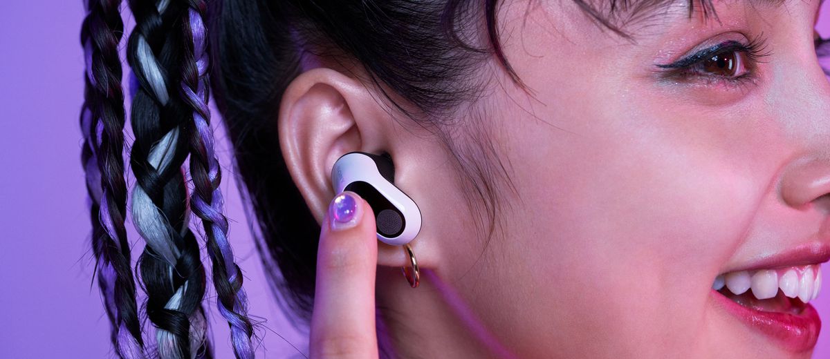 Une photo prise par Sony montrant une personne souriante portant ses écouteurs sans fil Inzone Buds.  Ils rapprochent un doigt de leur oreille pour régler un réglage sur les écouteurs.