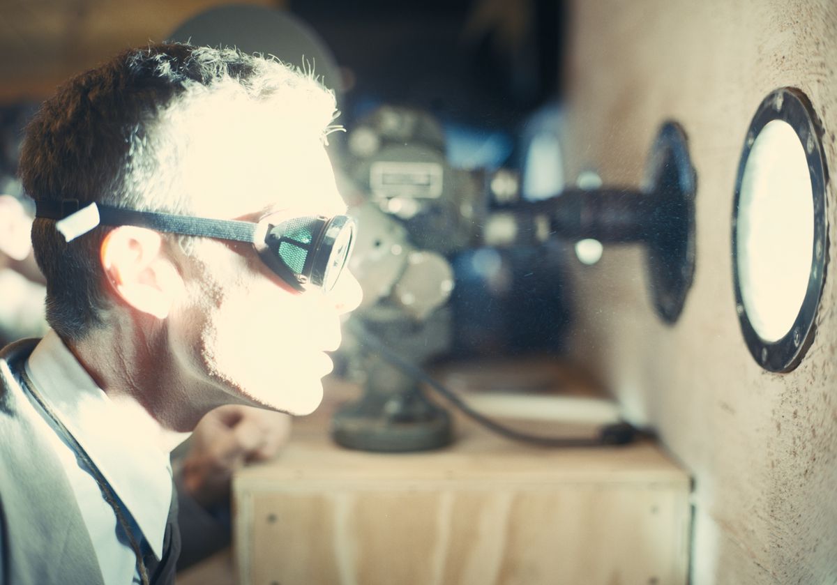 Dans le rôle de J. Robert Oppenheimer dans le film Oppenheimer, Cillian Murphy porte des lunettes et regarde à travers un hublot une explosion projetant une lumière intensément brillante sur son visage.