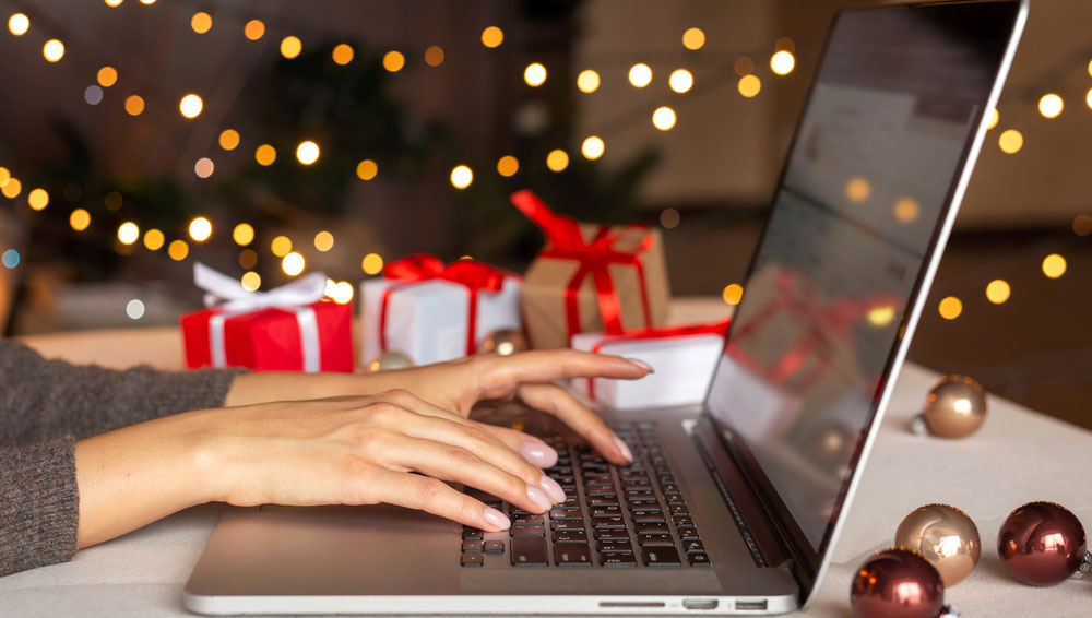 Les mains d'une femme tapent sur un MacBook avec un arbre de Noël et des décorations de Noël tout autour.