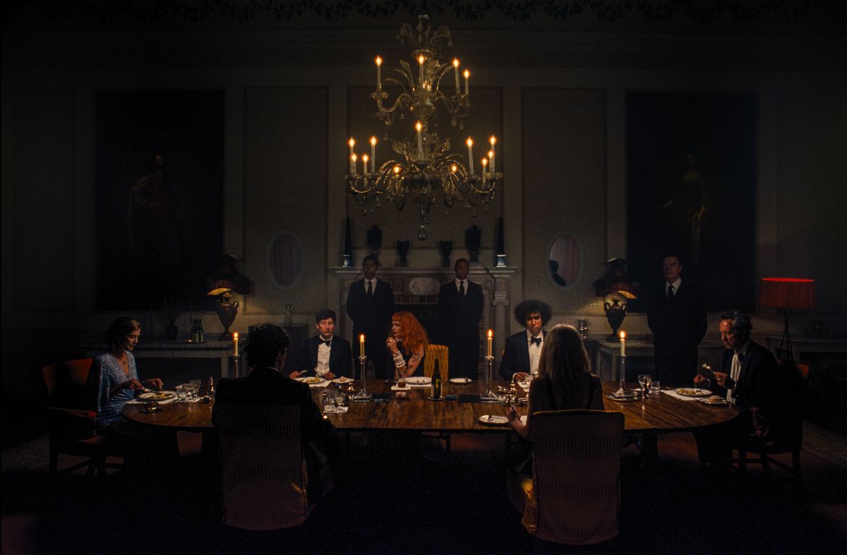 Oliver (Barry Keoghan) est assis à une longue table de salle à manger dans une pièce très sombre avec Felix (Jacob Jacob Elordi) et d'autres membres de sa famille, tous en tenue de soirée, dans le film Saltburn