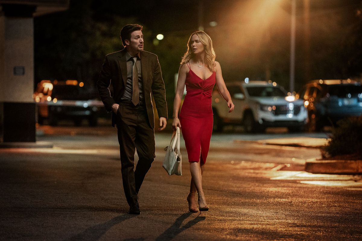 Emily Blunt, vêtue d'une robe rouge vif, marche à côté de Chris Evans, en costume sombre, dans un parking la nuit