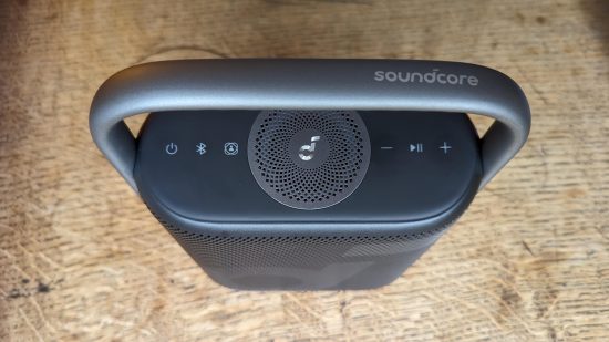 Test Anker Soundcore Motion X500 : un haut-parleur noir avec poignée argentée apparaît d'en haut sur une surface en bois.