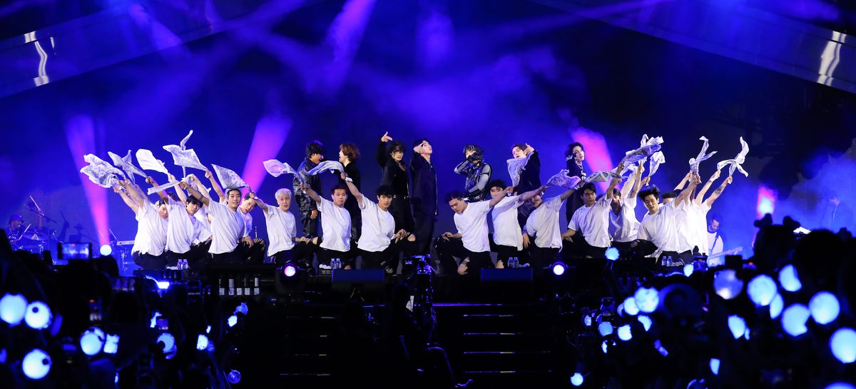 Les membres de BTS, sur scène pendant le concert Yet to Come, derrière une rangée de danseurs agenouillés en pantalons noirs et T-shirts blancs, agitant des vêtements blancs.