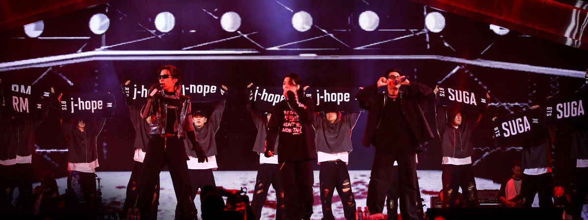 Les membres de BTS sur scène lors du concert Yet to Come, mélangés à des danseurs en sweat à capuche gris brandissant des pancartes indiquant « j-hope », « j-nope » et « SUGA ».