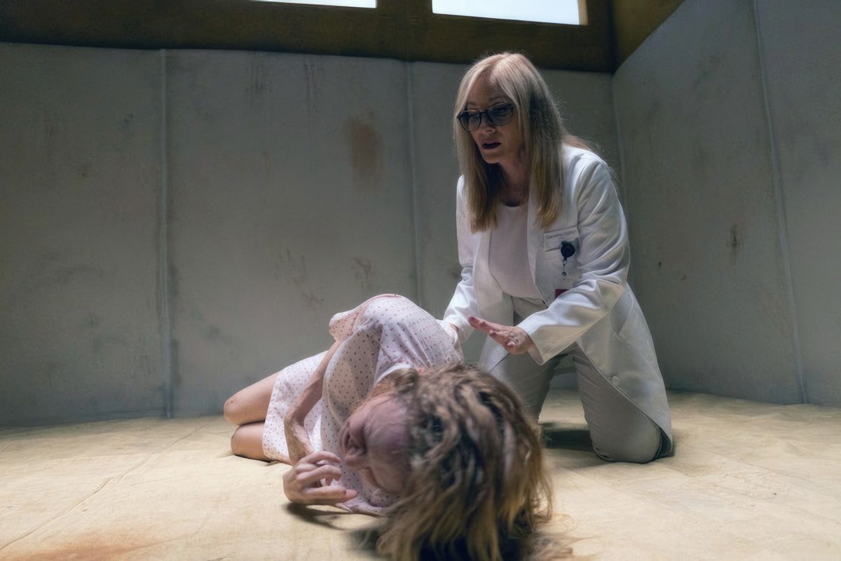 Elizabeth (Heather Graham, vêtue d'une blouse d'hôpital), se recroqueville en pleurant sur le sol d'une cellule nue d'un hôpital psychiatrique tandis qu'une amie psychiatre (Barbara Crampton) s'agenouille à côté d'elle et lui tend la main pour la soutenir dans Adapted Flesh.