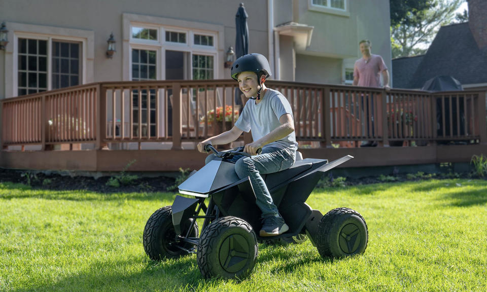 Photo marketing de style de vie d'un garçon chevauchant joyeusement un VTT Tesla Cyberquad for Kids.  Son père rayonne de dos alors qu'il se tient sur le porche ensoleillé de banlieue de la famille.