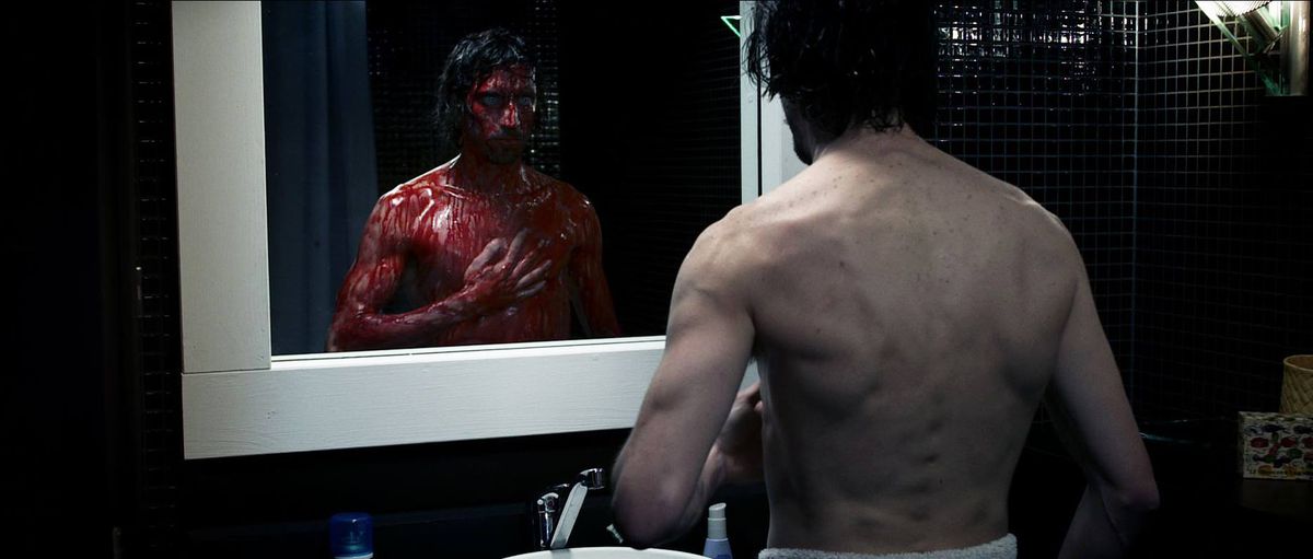Un homme se tient devant le miroir de sa salle de bain et vérifie sa poitrine couverte de sang dans Chimères.