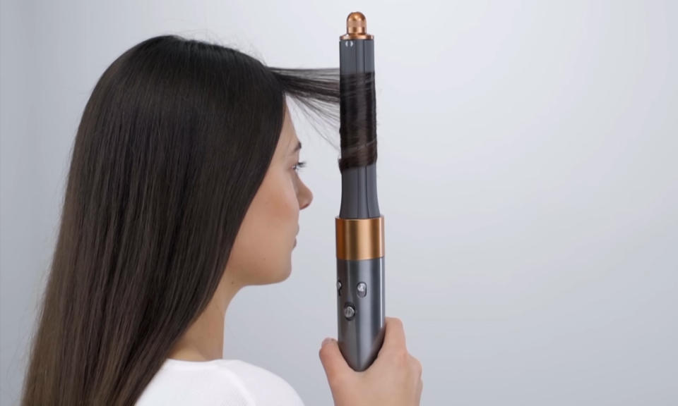 Image tirée d'une vidéo de produit montrant une femme tenant verticalement l'outil de coiffure Dyson Airwrap alors qu'il enroule ses longs cheveux noirs autour de son corps.  Fond gris.